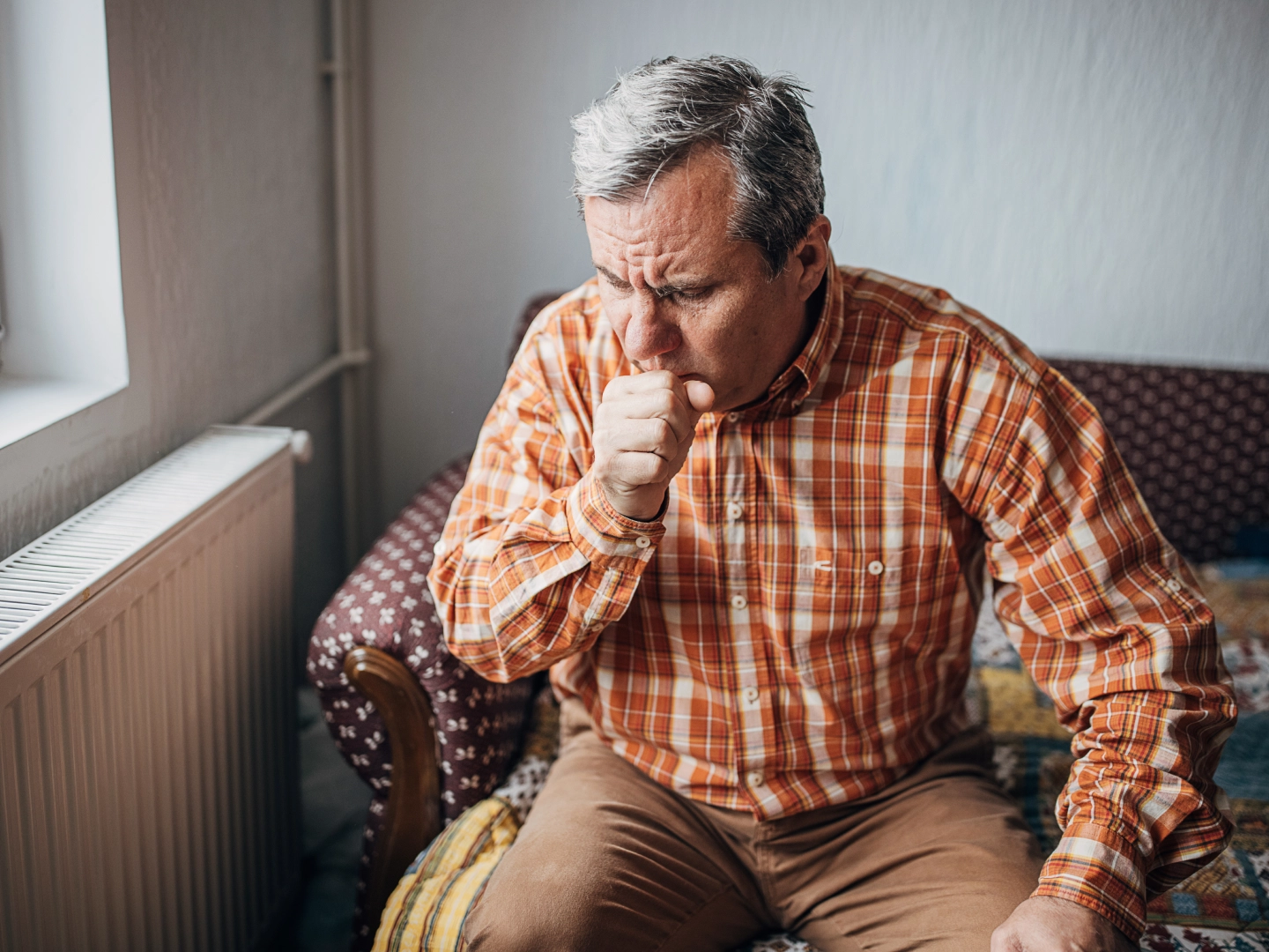 Older man sitting on chair near window coughing.