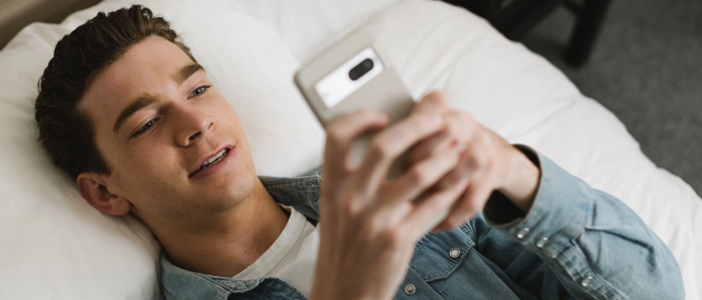 Young man in bed using his mobile phone to take a mental health visit online.